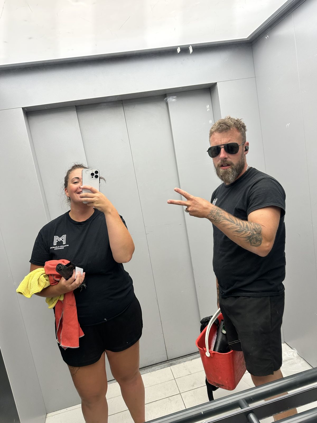 Rhianna and Chris in Mackies t-shirts taking a mirror selfie in a lift between jobs, cloths and a red bucket of kit in hand
