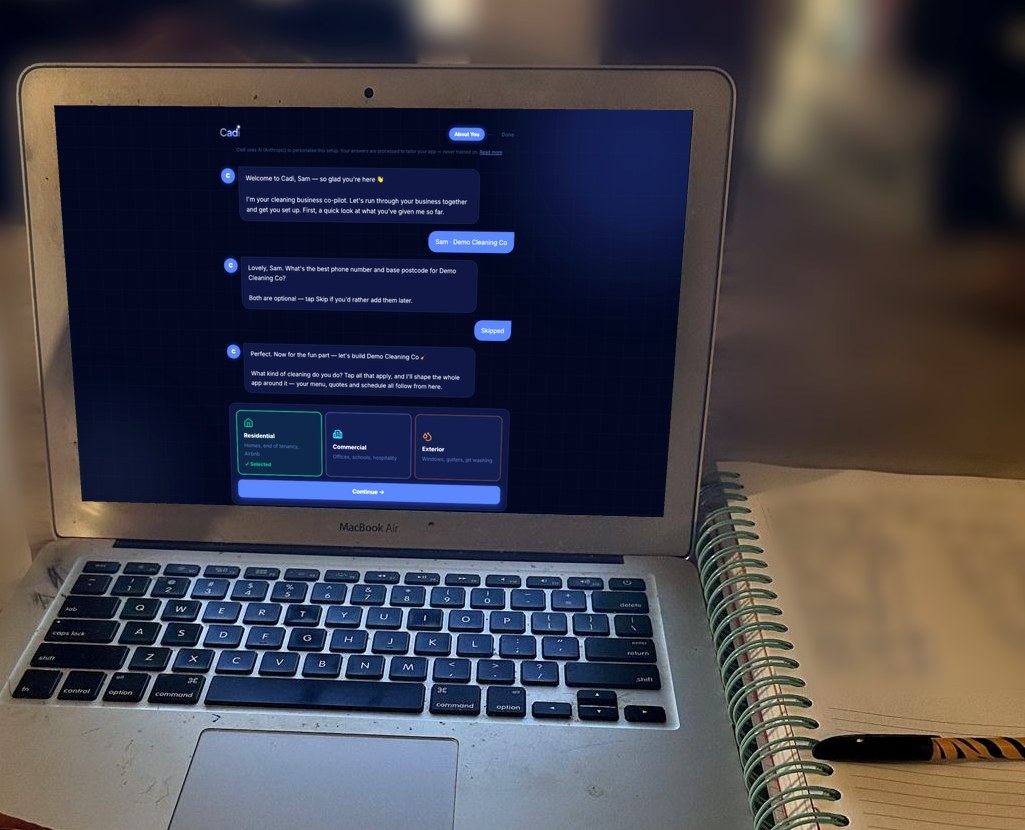A laptop at the kitchen table late at night showing Cadi’s setup chat — Cadi asks what kind of cleaning the business does — next to a spiral notebook with a handwritten to-do list